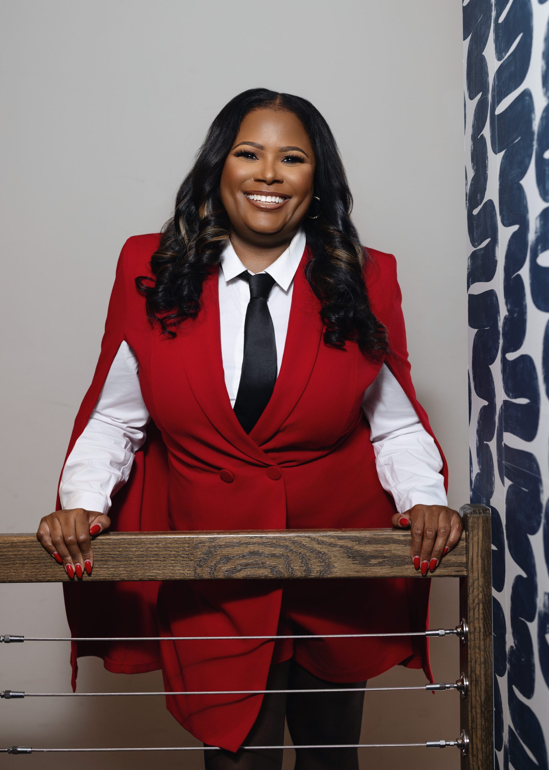 Professional portrait of a confident female real estate agent wearing a red blazer, smiling while leaning on a modern wooden railing.